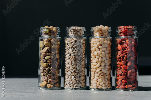 Set of glass bottles with pistachio, sunflower seeds, pine nuts and goji berry isolated on black background. Assorted healthy products in containers
