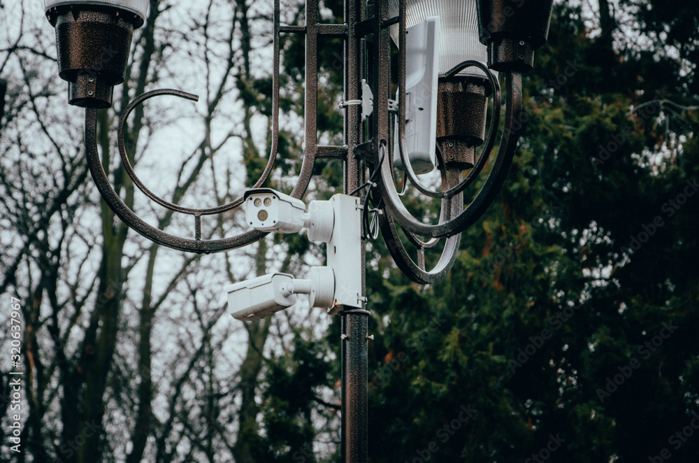 White surveillance camera hanging on a pole. The camera watches the order in the streets and in the parks. Camera on a post in a park. Side view and straight White surveillance camera hanging on a pole. The camera watches the order in the streets and in the parks. Camera on a post in a park. Side view and straight