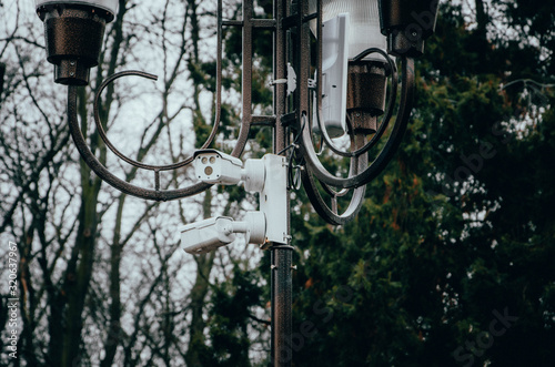 White surveillance camera hanging on a pole. The camera watches the order in the streets and in the parks. Camera on a post in a park. Side view and straight