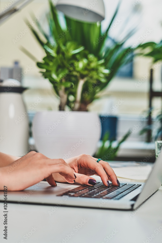 Girl with manicure is typing on a laptop, there are green plants on the background.