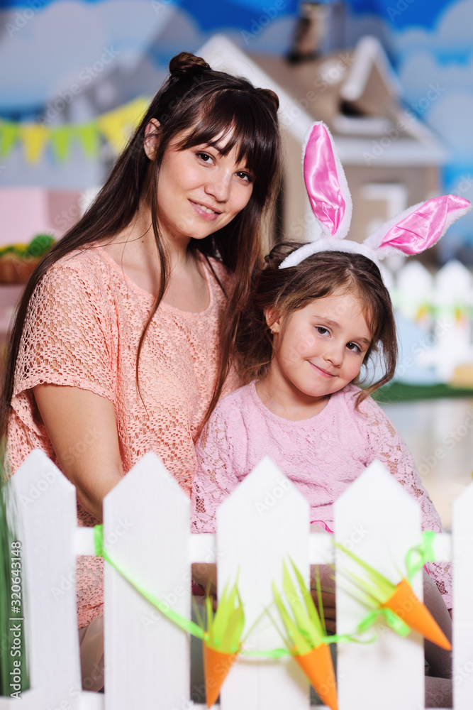 Obraz premium Mom with daughter with rabbit ears smiling sitting on white fence background. Celebrating Easter