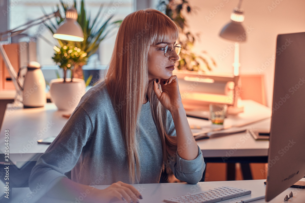 In the nice office blond woman in glasses is focused on computer while doing her job.