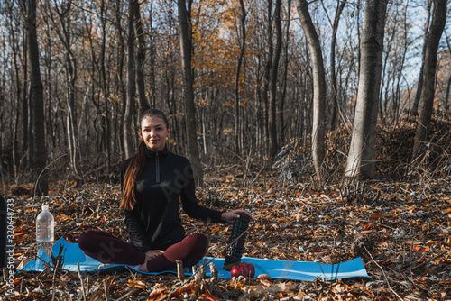 Young attractive slim brunette girl sitting on the blue yoga mat among the forest, holds black roller in her hand and looking at the camera, bottle with water stands deside photo