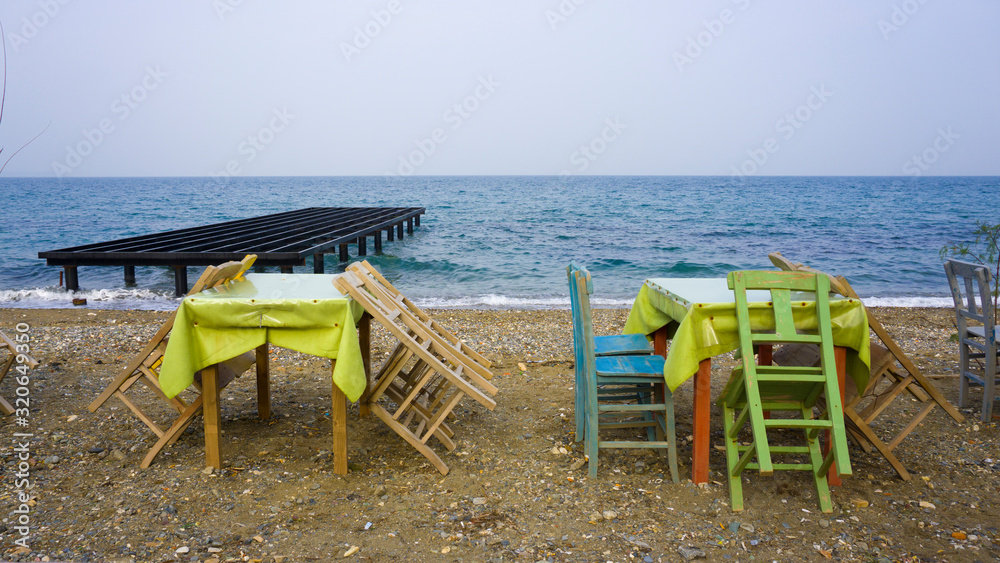 Empty wooden chairs and tables lined up on pebble beach. Seaside restaurant ready for sunny weekend customers. Relaxing place for evening or night meal. Outdoor resort cafe for eating or drinking.