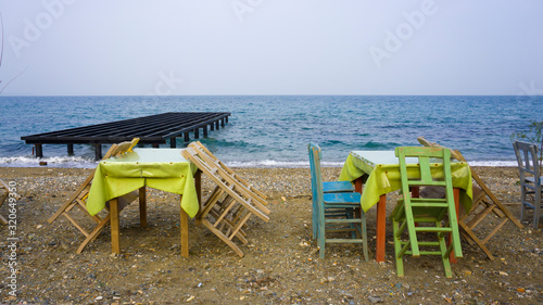 Empty wooden chairs and tables lined up on pebble beach. Seaside restaurant ready for sunny weekend customers. Relaxing place for evening or night meal. Outdoor resort cafe for eating or drinking. © Akin Ozcan