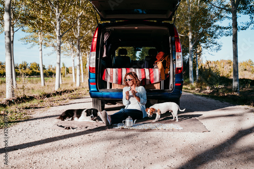 young beautiful woman drinking coffee or tea camping outdoors with a van and her two dogs. Travel concept