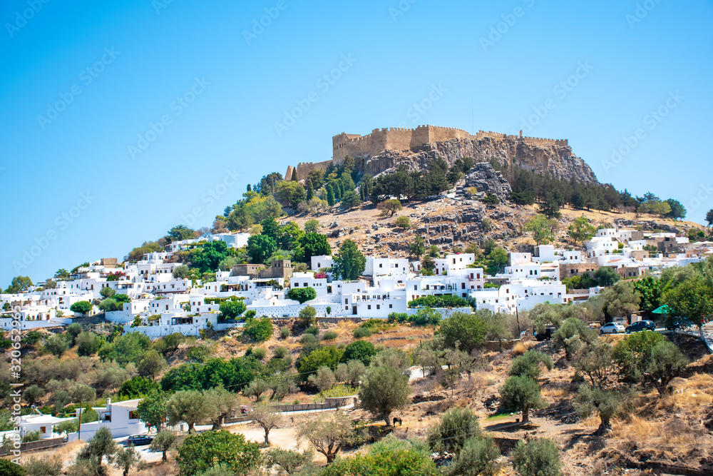 Obraz premium View of white houses of village of Lindos among trees with Acropolis in background (Rhodes, Greece)