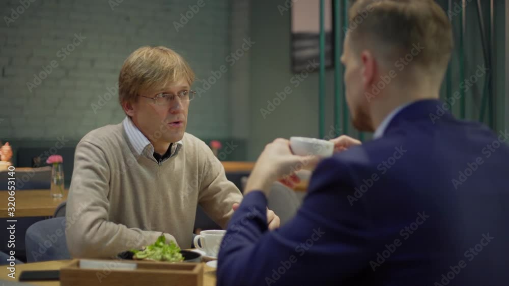 Tracking right medium shot of middle aged businessman in eyeglasses talking to business partner over coffee and breakfast during meeting at cafe table Tracking right medium shot of middle aged businessman in eyeglasses talking to business partner over coffee and breakfast during meeting at cafe table