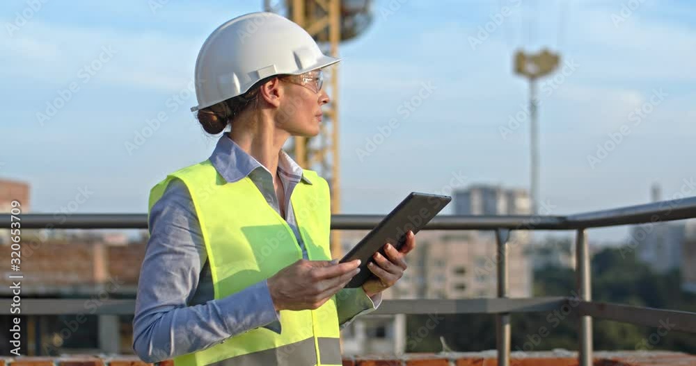 Caucasian young beautiful woman architect or builder in the hardhat standing at the roof of the building site and using tablet device, tapping and scrolling. Caucasian young beautiful woman architect or builder in the hardhat standing at the roof of the building site and using tablet device, tapping and scrolling.