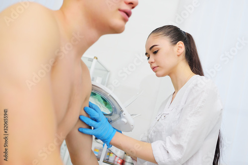 Fototapeta Naklejka Na Ścianę i Meble -  doctor dermatologist-a young woman examines the skin condition of a male patient-moles, scars, pigmentation through a special device-a cosmetic lamp magnifying glass. 