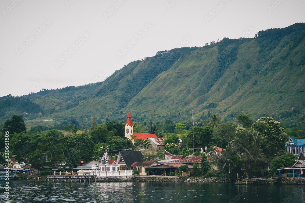 Volcanic Island of Samosir on the lake of Toba, on northern Sumatra, Visible typical houses and christian church on the island on a cloudy day. Volcanic Island of Samosir on the lake of Toba, on northern Sumatra, Visible typical houses and christian church on the island on a cloudy day.