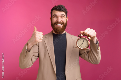 Happy young business man showing thumbs up gesture and holding alarm clock over pink background photo