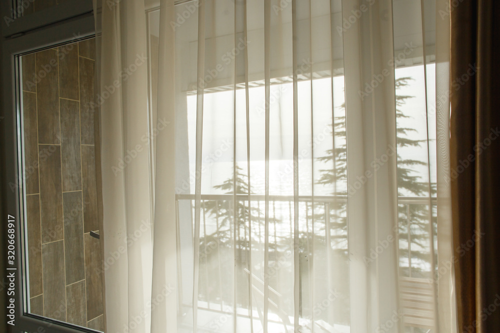 Daylight shining through the white transparent curtain. Beautiful view from the window on the trees and the sea