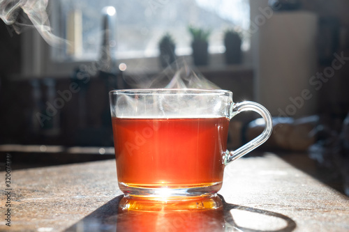 Hot aromatic tea in glass cup with steam on the kitchen table on sunlight