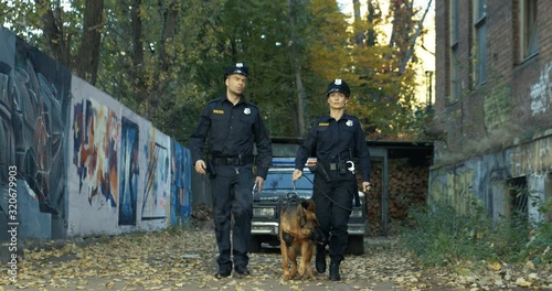 Caucasian policeman and policewoman in uniforms walking with shepherd dog. Male and female police officers searching something and patrolling disrict. photo