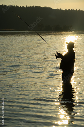 Fishing in the early morning on the lake in the summer. Silhouette of the fisherman with fishing rod in hand in front of morning sunlight. In the distance - a dark forest. With copy space