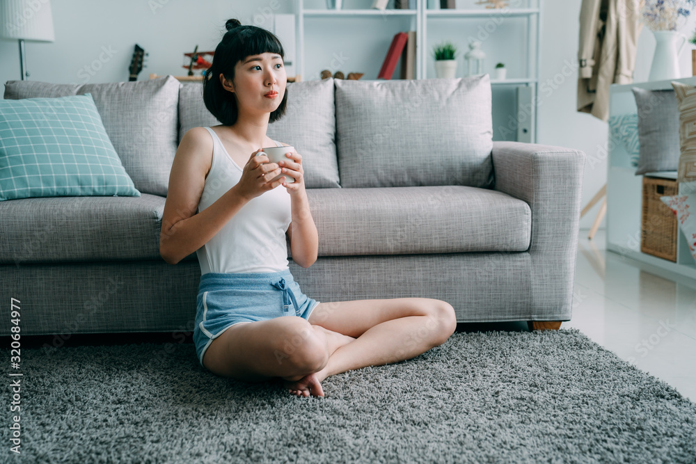 Peaceful woman relaxing at home with cup of tea in bright morning. asian chinese girl sitting on carpet wearing sleepwear holding coffee to awake. lady leaning on comfort couch in house living room Peaceful woman relaxing at home with cup of tea in bright morning. asian chinese girl sitting on carpet wearing sleepwear holding coffee to awake. lady leaning on comfort couch in house living room