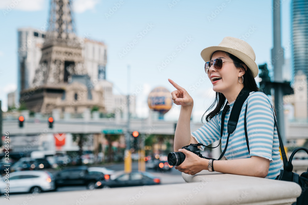Young woman tourist pointing aside showing view with curiously smiling face. girl photographer holding camera sightseeing busy modern city in summer holidays usa. famous hotel and tower in las vegas