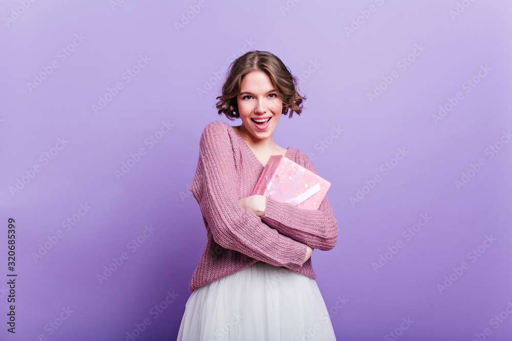 Attractive girl with curly hairstyle holding new year present and smiling to camera. Studio shot of elegant woman in white skirt posing with pink gift box on purple background. Attractive girl with curly hairstyle holding new year present and smiling to camera. Studio shot of elegant woman in white skirt posing with pink gift box on purple background.