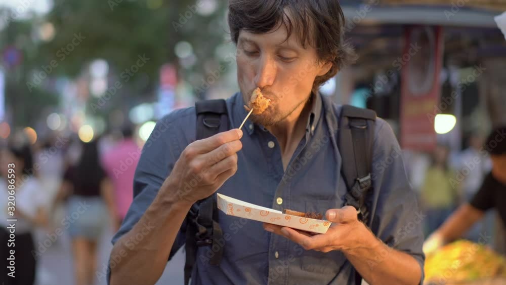Young man visits the traditional korean street market. He is eating fish cake on a bamboo stick. Travel to Korea.