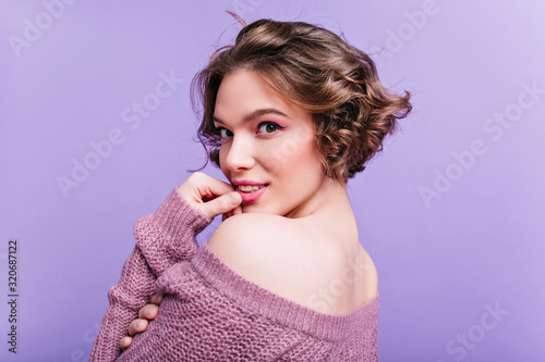 Close-up portrait of interested young woman with short brunette hair wears sweater. Indoor photo of sensual lady playfully looking over shoulder on purple background.