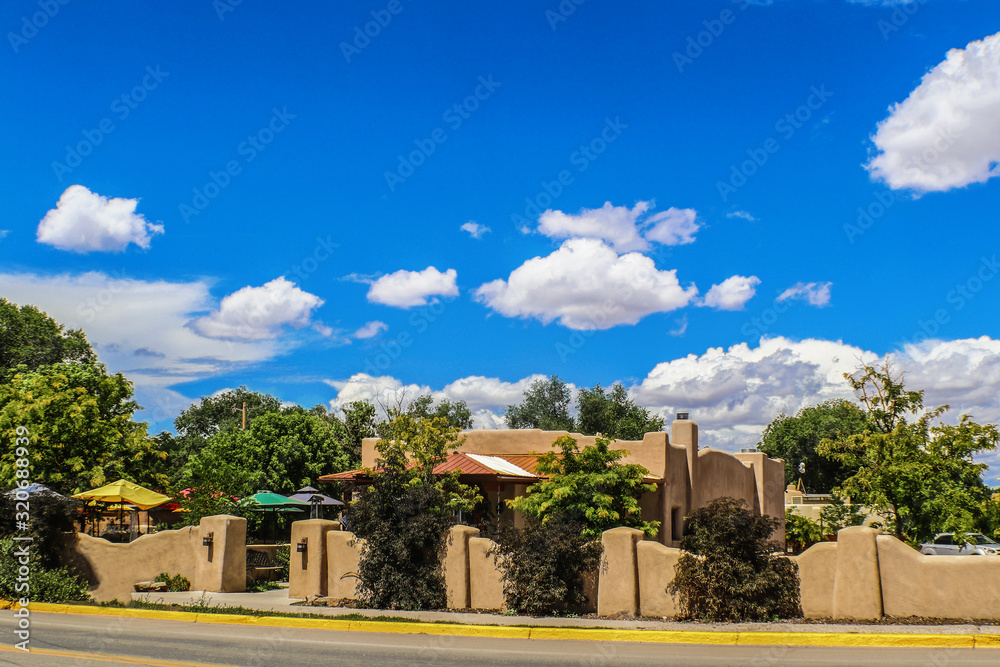 Beautiful stucco mud southwestern USA house with fence and landscaping and umbrellas beside road with curb painted yellow under very blue sky with puffy clouds