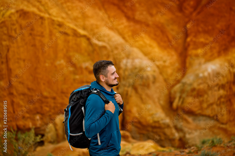 A young male tourist with a backpack stands on top of a mountain. Red mountains in the Altai.Close-up portrait. Traveler in the mountains.Outdoor activity.Summer adventure travel. A young male tourist with a backpack stands on top of a mountain. Red mountains in the Altai.Close-up portrait. Traveler in the mountains.Outdoor activity.Summer adventure travel.