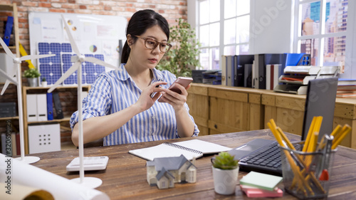Contemporary construction worker with electronic equipment. young lady engineer looking at cellphone and texting message online discussing with colleague. woman architect in office using mobile phone