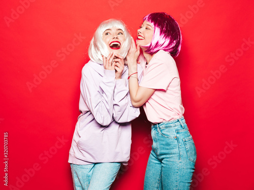 Two young sexy hipster girls in wigs and red lips.Beautiful trendy women in summer clothes.Carefree models posing near red wall in studio.Positive female going crazy