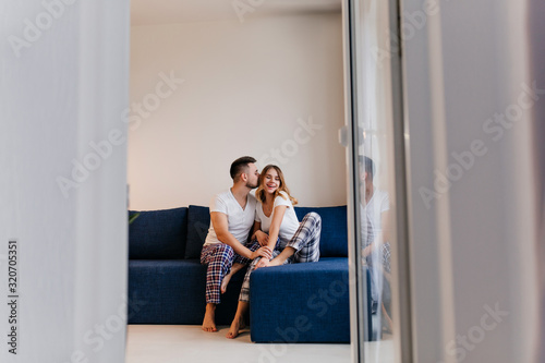Funny barefooted man in pajama kissing his wife. Indoor portrait of lazy married couple enjoying morning.