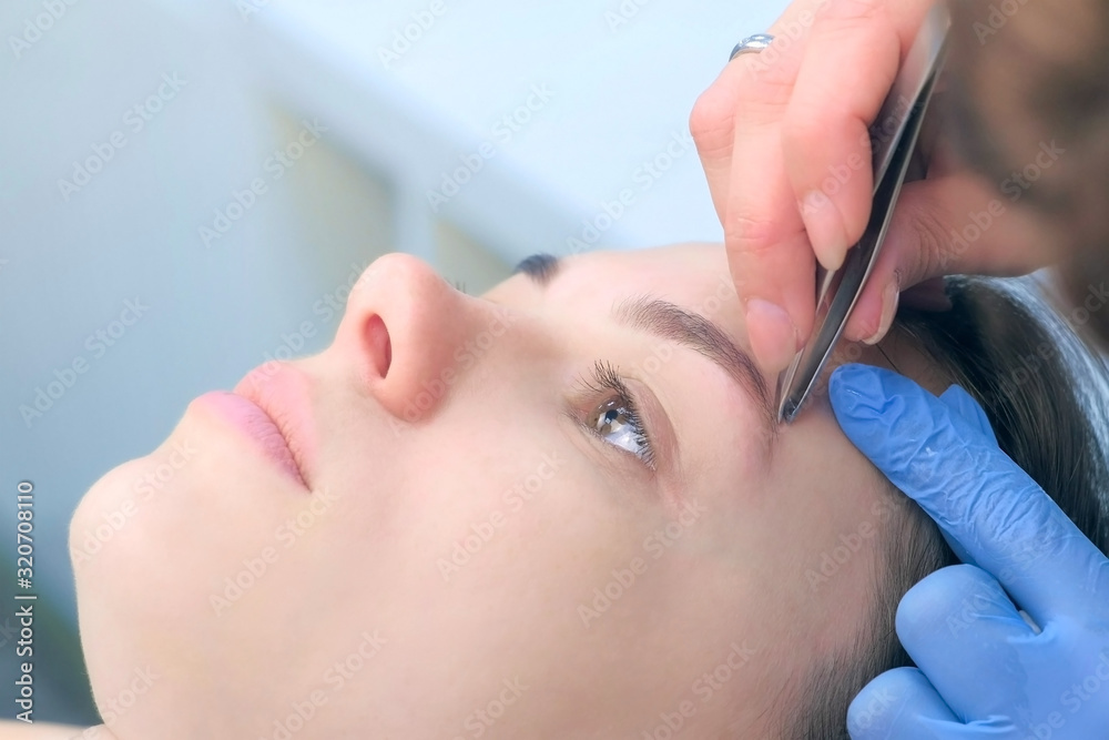 Professional beautician plucking eyebrows with tweezers to woman in beauty salon after tint eyebrow procedure, closeup view. Cosmetologist shaping girl's eyebrows in cosmetology clinic. Professional beautician plucking eyebrows with tweezers to woman in beauty salon after tint eyebrow procedure, closeup view. Cosmetologist shaping girl's eyebrows in cosmetology clinic.