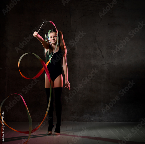 Young smiling girl gymnast in black sport body and uppers standing and making exercise with colorful gymnastic tape