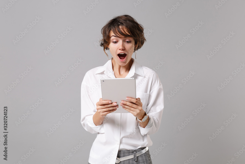 Shocked young business woman in white shirt posing isolated on grey wall background studio portrait. Achievement career wealth business concept. Mock up copy space. Holding, using tablet pc computer. Shocked young business woman in white shirt posing isolated on grey wall background studio portrait. Achievement career wealth business concept. Mock up copy space. Holding, using tablet pc computer.