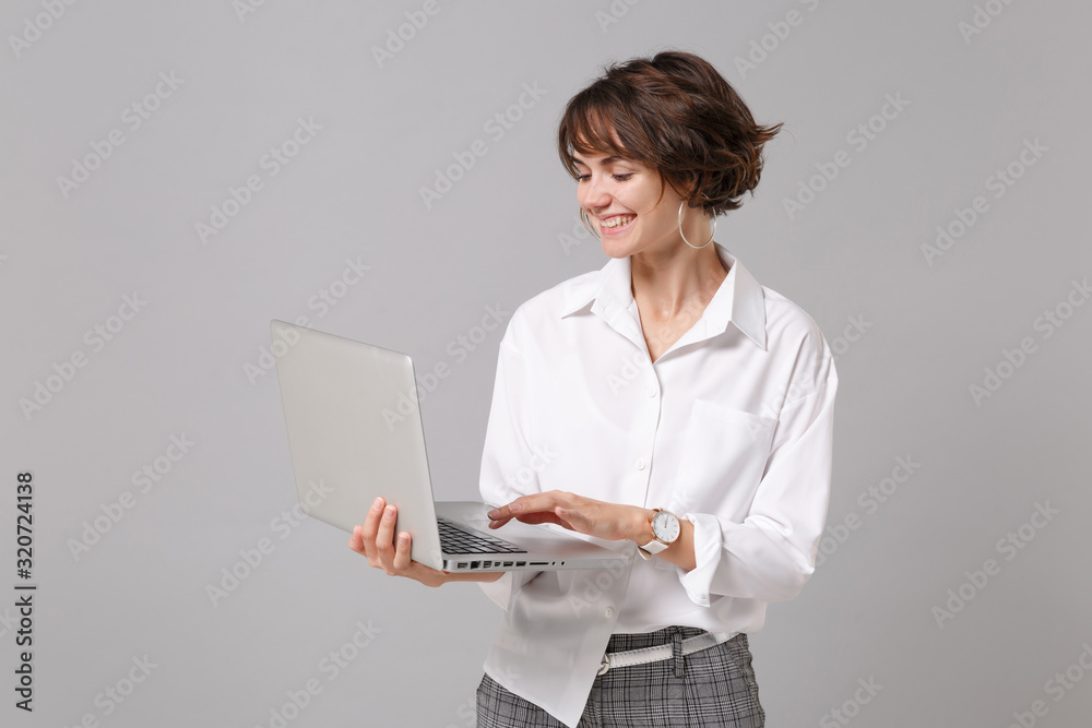 Smiling young business woman in white shirt posing isolated on grey wall background studio portrait. Achievement career wealth business concept. Mock up copy space. Working on laptop pc computer. Smiling young business woman in white shirt posing isolated on grey wall background studio portrait. Achievement career wealth business concept. Mock up copy space. Working on laptop pc computer.