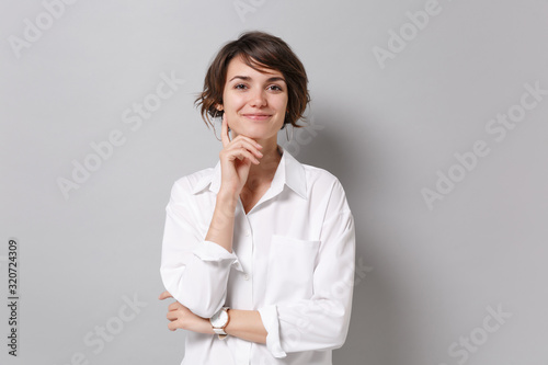 Smiling young business woman in white shirt posing isolated on grey background studio portrait. Achievement career wealth business concept. Mock up copy space. Put hand prop up on chin looking camera.