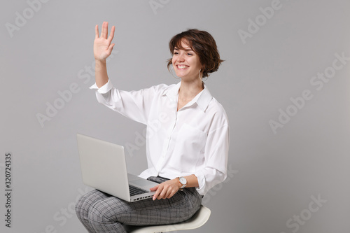 Pleasant young business woman in white shirt isolated on grey background. Achievement career wealth business concept. Hold laptop pc computer, sitting, waving greeting with hand as notices someone.