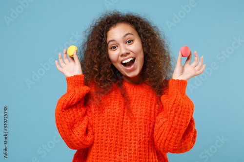 Excited young african american girl in casual orange knitted clothes isolated on pastel blue background studio portrait. People emotions lifestyle concept. Mock up copy space. Hold french macarons. © ViDi Studio