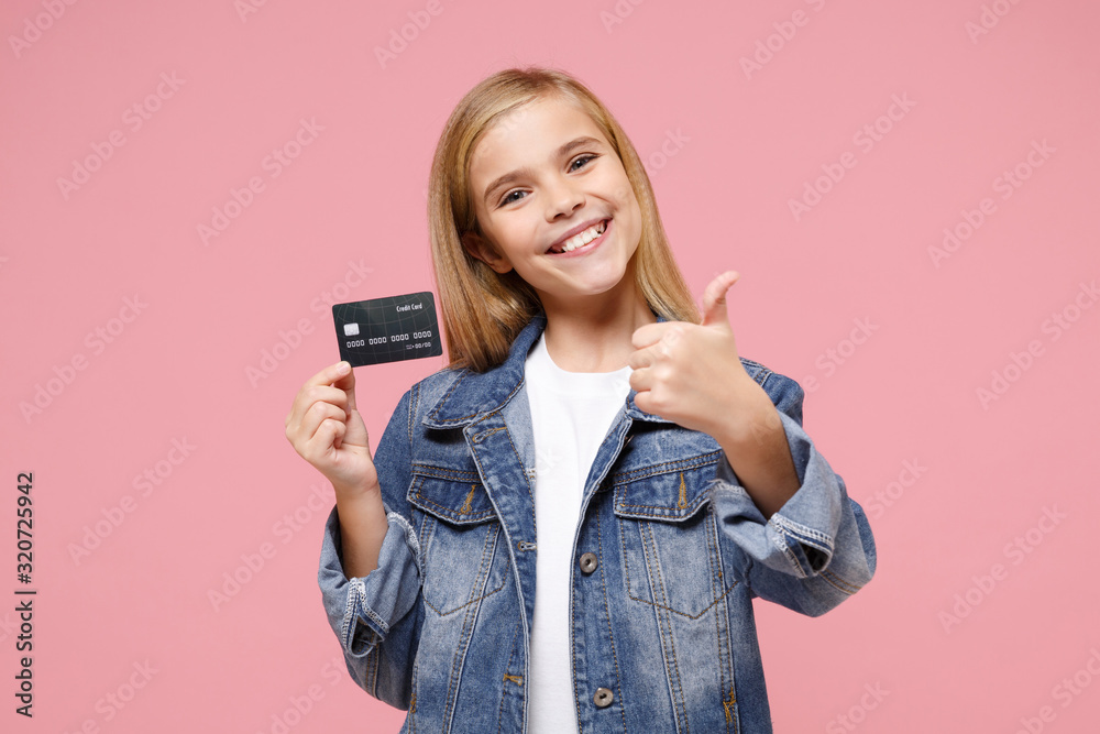 Smiling little blonde kid girl 12-13 years old in denim jacket posing isolated on pastel pink wall background. Childhood lifestyle concept. Mock up copy space. Showing thumb up, hold credit bank card. - obrazy, fototapety, plakaty 