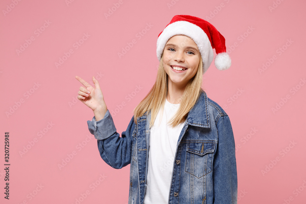 Smiling little blonde kid girl 12-13 years old in denim jacket Christmas hat isolated on pastel pink background. Happy New Year 2020 celebration concept. Mock up copy space. Point index finger aside. Smiling little blonde kid girl 12-13 years old in denim jacket Christmas hat isolated on pastel pink background. Happy New Year 2020 celebration concept. Mock up copy space. Point index finger aside.