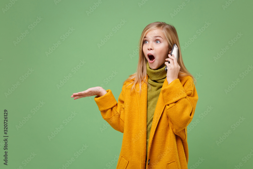 Shocked little blonde kid girl 12-13 years old in yellow coat posing isolated on pastel green background. Childhood lifestyle concept. Mock up copy space. Talking on mobile phone, spreading hands. Shocked little blonde kid girl 12-13 years old in yellow coat posing isolated on pastel green background. Childhood lifestyle concept. Mock up copy space. Talking on mobile phone, spreading hands.