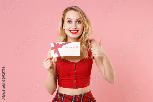 Smiling young blonde woman girl in red sexy clothes isolated on pastel pink background studio portrait. People emotions lifestyle concept. Mock up copy space. Hold gift certificate, showing thumb up.