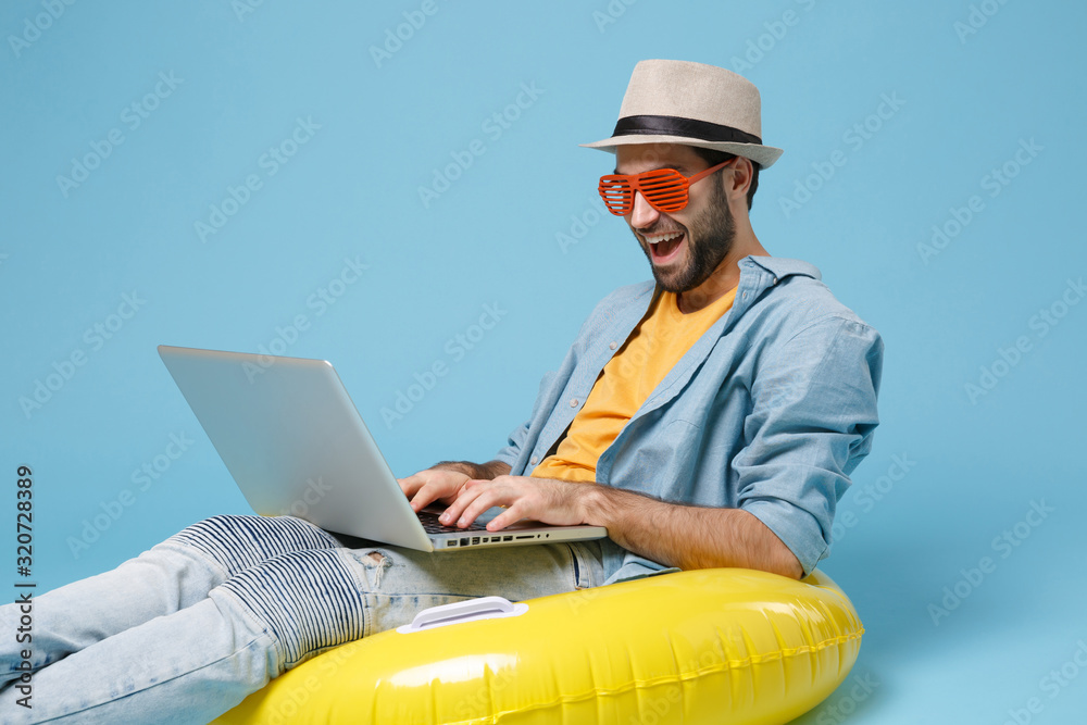 Cheerful traveler tourist man in yellow clothes glasses isolated on blue background. Passenger traveling abroad on weekend. Air flight journey concept. Sit in rubber ring work on laptop booking hotel.