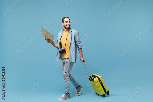 Cheerful traveler tourist man in yellow casual clothes with photo camera isolated on blue background. Male passenger traveling abroad on weekends. Air flight journey concept. Hold suitcase, city map.