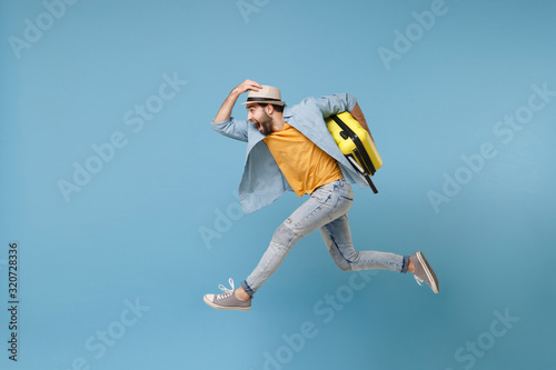 Side view of excited traveler tourist man in yellow clothes isolated on blue background. Male passenger traveling abroad on weekends. Air flight journey concept. Jumping like running, hold suitcase.