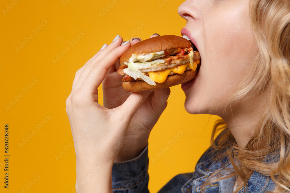 Cropped image side view of young woman in denim clothes isolated on orange background studio portrait. Proper nutrition or American classic fast food concept. Mock up copy space. Eating biting burger. Cropped image side view of young woman in denim clothes isolated on orange background studio portrait. Proper nutrition or American classic fast food concept. Mock up copy space. Eating biting burger.