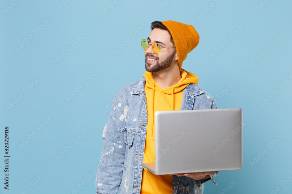 Smiling young hipster guy in fashion jeans denim clothes posing isolated on pastel blue background in studio. People lifestyle concept. Mock up copy space. Holding laptop pc computer, looking aside.