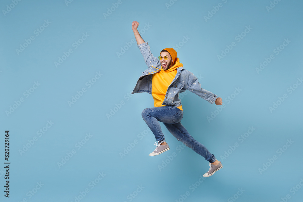 Crazy young hipster guy in fashion jeans denim clothes posing isolated on pastel blue background. People lifestyle concept. Mock up copy space. Jumping, spreading hands, clenching fists like winner.