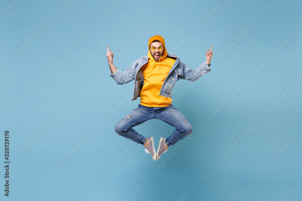 Crazy young hipster guy in fashion jeans denim clothes posing isolated on pastel blue wall background studio portrait. People emotions lifestyle concept. Mock up copy space. Jumping showing horns up.