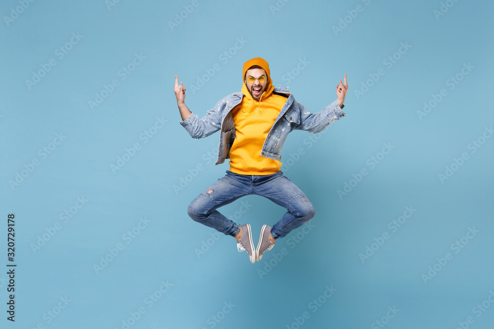Crazy young hipster guy in fashion jeans denim clothes posing isolated on pastel blue wall background studio portrait. People emotions lifestyle concept. Mock up copy space. Jumping showing horns up. Crazy young hipster guy in fashion jeans denim clothes posing isolated on pastel blue wall background studio portrait. People emotions lifestyle concept. Mock up copy space. Jumping showing horns up.
