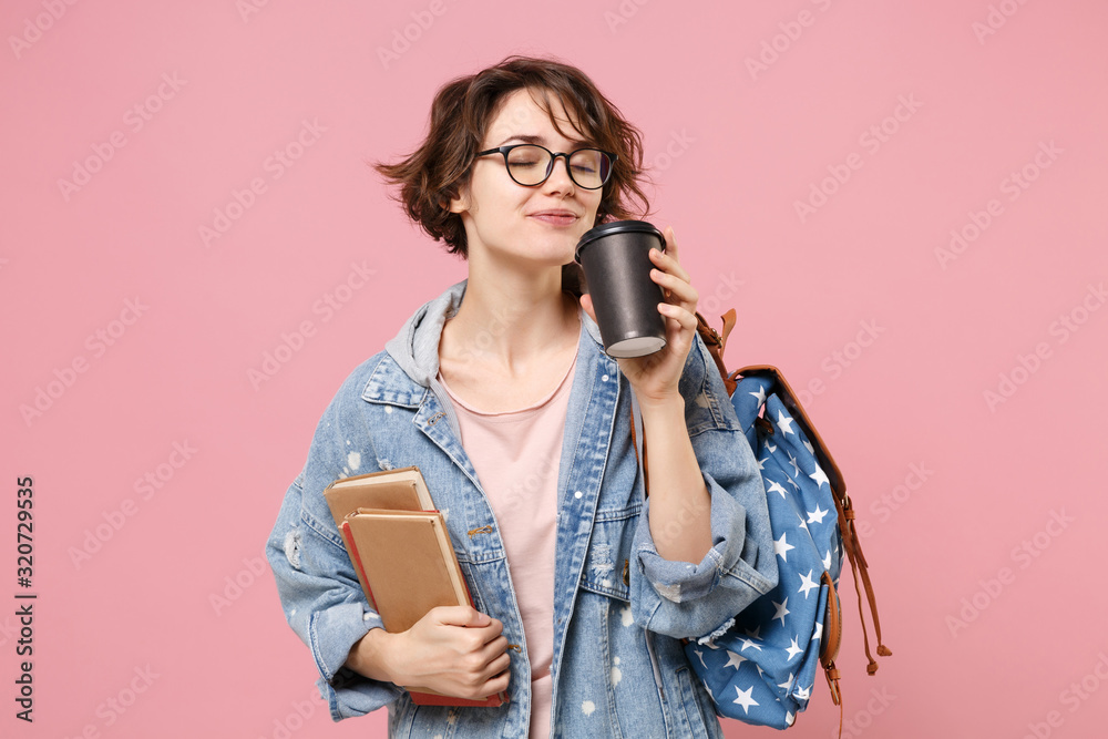 Pretty young woman student in denim clothes eyeglasses, backpack posing isolated on pastel pink background. Education in high school university college concept. Hold books paper cup of coffee or tea.